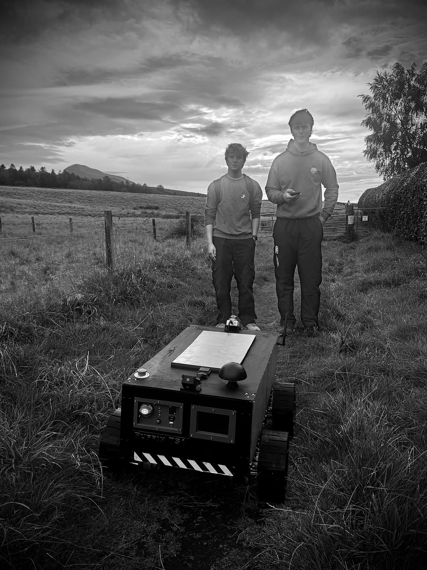 Two engineers standing in a field at dusk behind Puffin TS1, a tracked survey rover, with hills on the horizon.